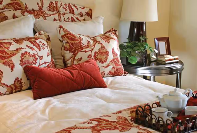 A neatly made bed with white bedding and decorative pillows featuring a red and beige floral pattern. A red rectangular pillow is placed in front. Next to the bed is a round dark wooden side table with a lamp, a small green plant, a picture frame, and a couple of books. A tea set with a teapot and cups is arranged on a tray on the bed.