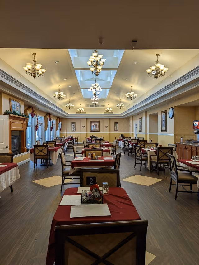 A spacious dining room with multiple tables covered in red and white tablecloths, each set with menus, condiments, and glasses. The room features elegant chandeliers hanging from a ceiling with skylights, large windows with curtains on the left side, framed artwork on the walls, and a fireplace on the left. The floor is wooden with light-colored square inlays, and there is a beverage dispenser on the right side.