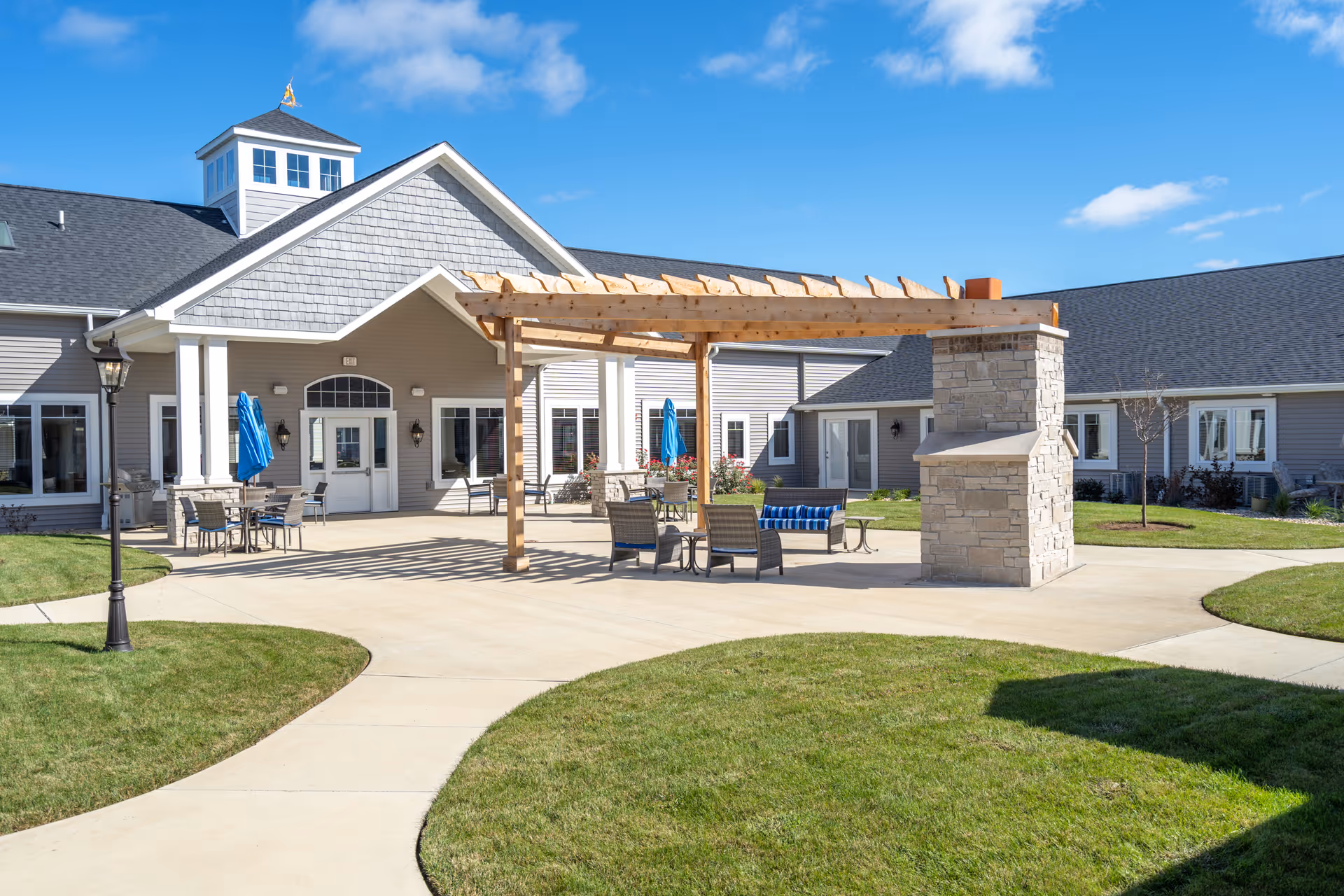 Outdoor patio area at Villas of Holly Brook Assisted Living & Memory Care in Bethalto, IL, featuring a wooden pergola with seating underneath, a stone fireplace, tables with umbrellas, and a well-maintained lawn and walkway leading to the building entrance.