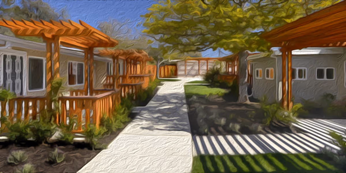 Outdoor walkway between single-story buildings with wooden pergolas and railings, surrounded by greenery and trees under a clear blue sky.