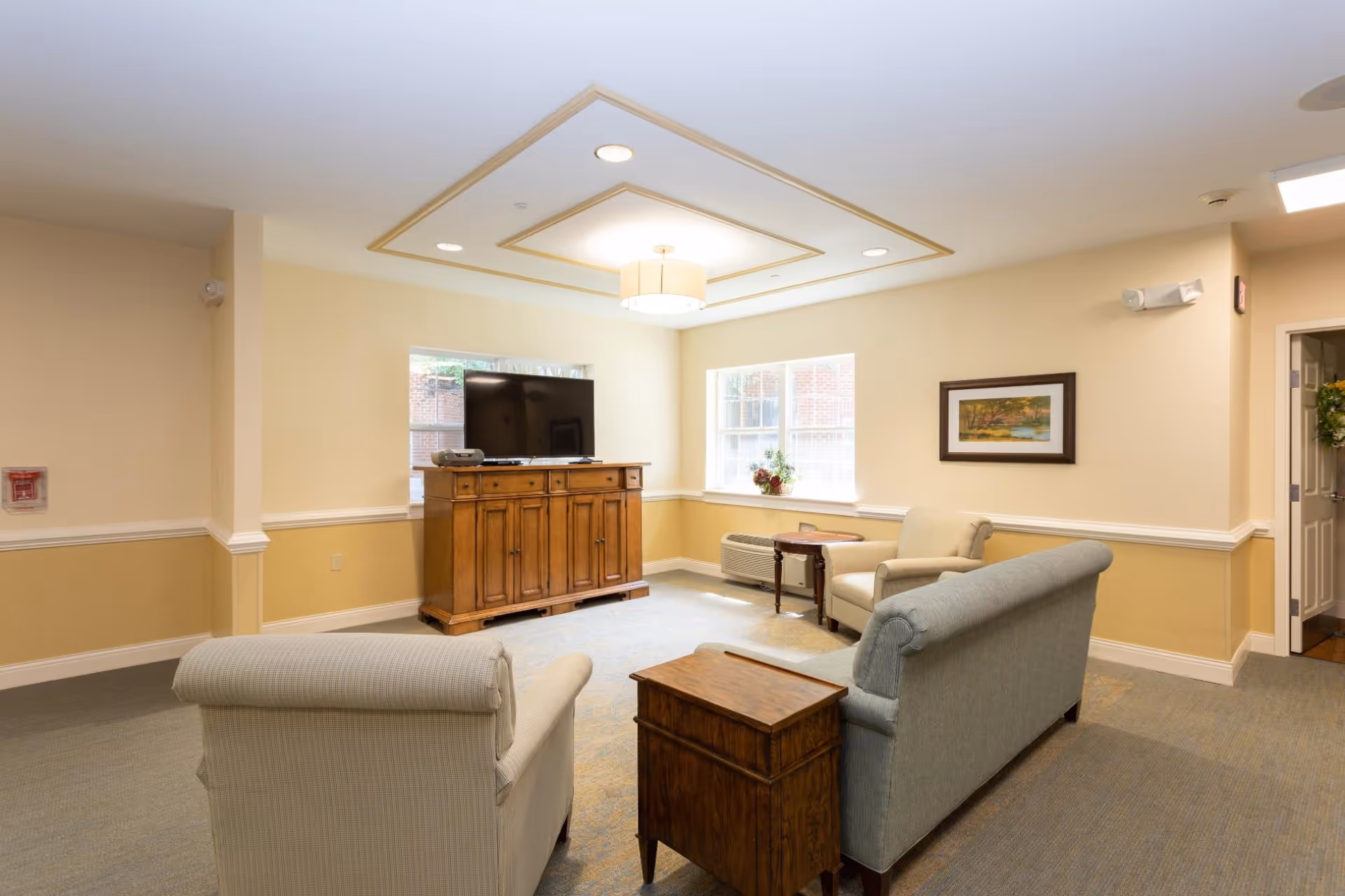A bright communal living room with upholstered sofas and chairs facing a wooden TV cabinet beneath windows.