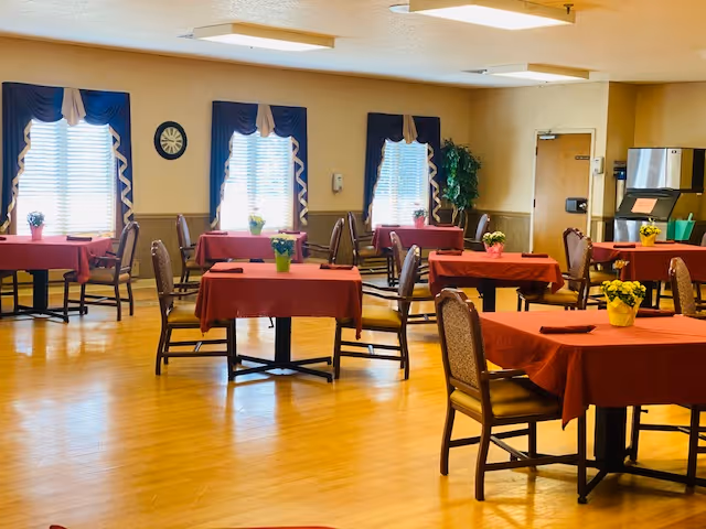 A dining room with several tables covered in red tablecloths, each table decorated with a small potted plant. The room has wooden floors, multiple windows with blue curtains, and chairs arranged around the tables. There is a water dispenser and some storage cabinets in the background.