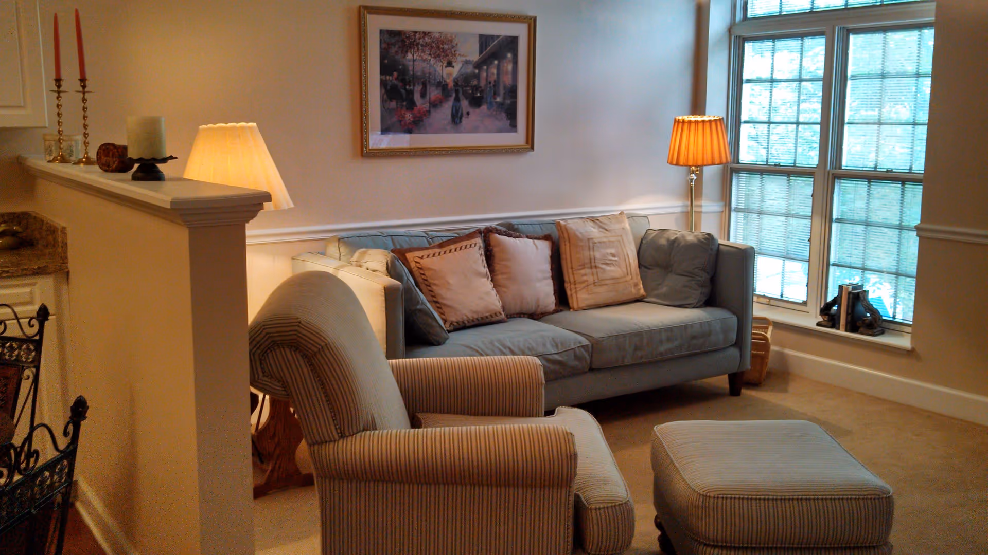 A cozy living room area with a light blue sofa adorned with four decorative pillows, a striped armchair with a matching ottoman, a floor lamp with a pleated shade, and a large window with blinds letting in natural light. A framed picture hangs on the wall above the sofa, and a half wall with candles and a lamp separates the living room from the adjacent space.
