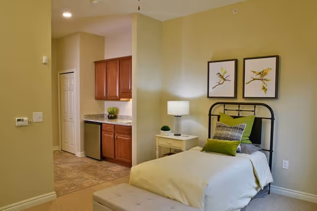 A cozy senior living bedroom featuring a single bed with green and gray pillows, a white nightstand with a lamp and small plant, two framed bird illustrations on the wall above the bed, and a kitchenette area with wooden cabinets and a small refrigerator in the background.