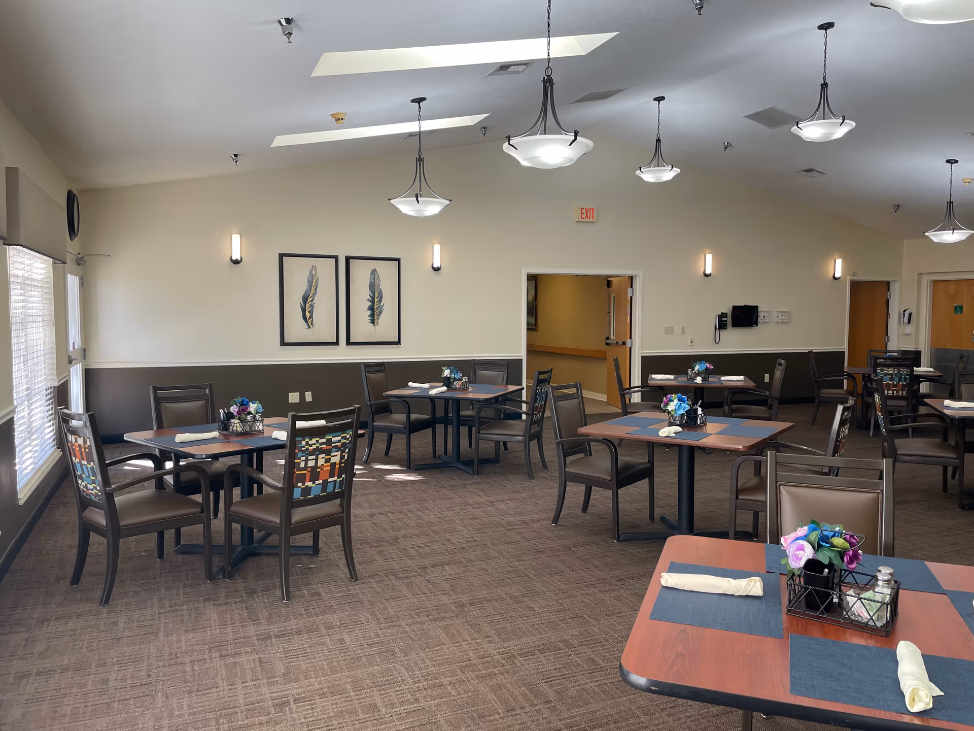 A dining room with several square wooden tables, each set with placemats, rolled napkins, and small flower arrangements. The room has beige walls with a darker wainscoting, carpeted floor, and multiple ceiling lights. There are framed feather artworks on the wall and windows with blinds on the left side.