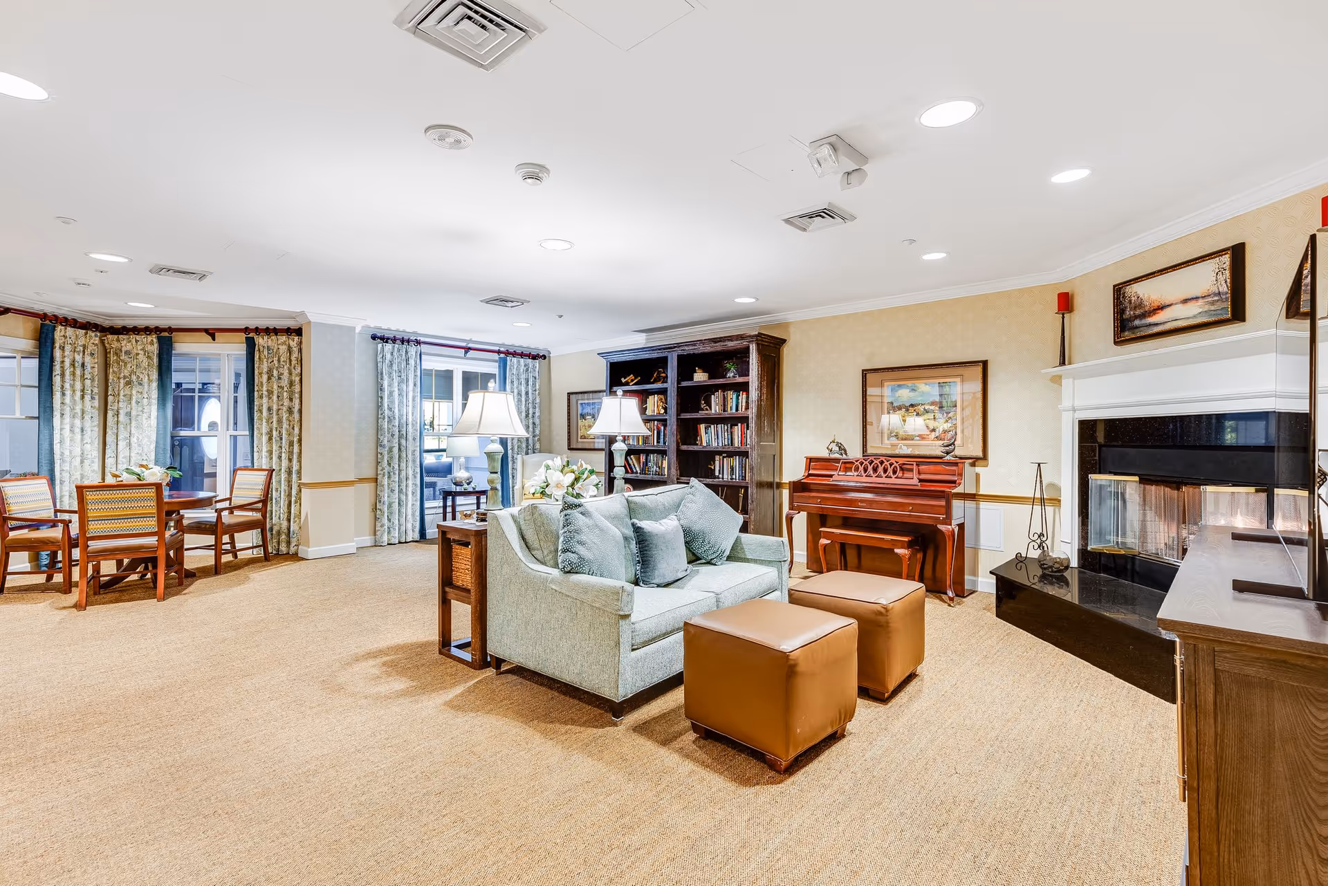 A bright and spacious living room area in a senior living facility featuring a light blue sofa with cushions, two brown ottomans, a wooden bookshelf filled with books, a wooden piano, a fireplace with a white mantel, framed artwork on the walls, and a dining table with chairs near large windows with patterned curtains.