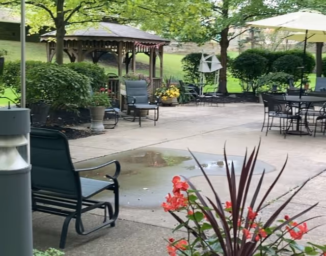 Outdoor patio area with metal chairs and tables, a gazebo, green trees, bushes, and flowering plants. There is a small puddle on the concrete ground and an umbrella providing shade over one of the tables.