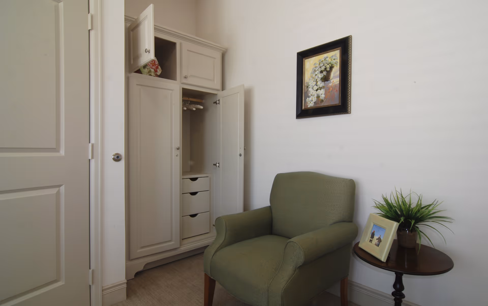 A small room with a green upholstered armchair next to a round wooden side table holding a potted plant and a framed photo. Behind the chair is a white built-in wardrobe with open doors revealing hanging space and drawers. A framed painting of white flowers hangs on the wall above the chair.