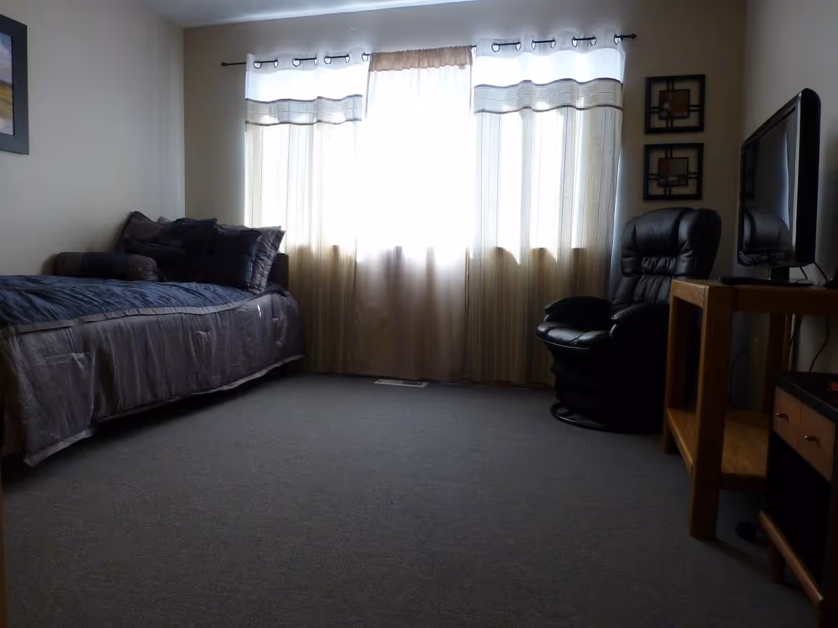 A bedroom with a bed on the left side covered with dark bedding and multiple pillows. A large window with sheer curtains lets in bright light in the center. On the right side, there is a black leather recliner chair next to a wooden TV stand holding a flat-screen television. The room has carpeted flooring and neutral-colored walls with two framed artworks hanging above the chair.
