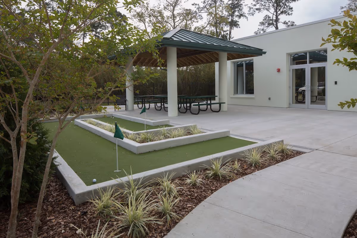 Outdoor area at Mount Carmel Gardens featuring a small putting green with three holes marked by green flags, surrounded by landscaping with plants and mulch. There is a covered pavilion with picnic tables and a building with large windows and a glass door in the background.