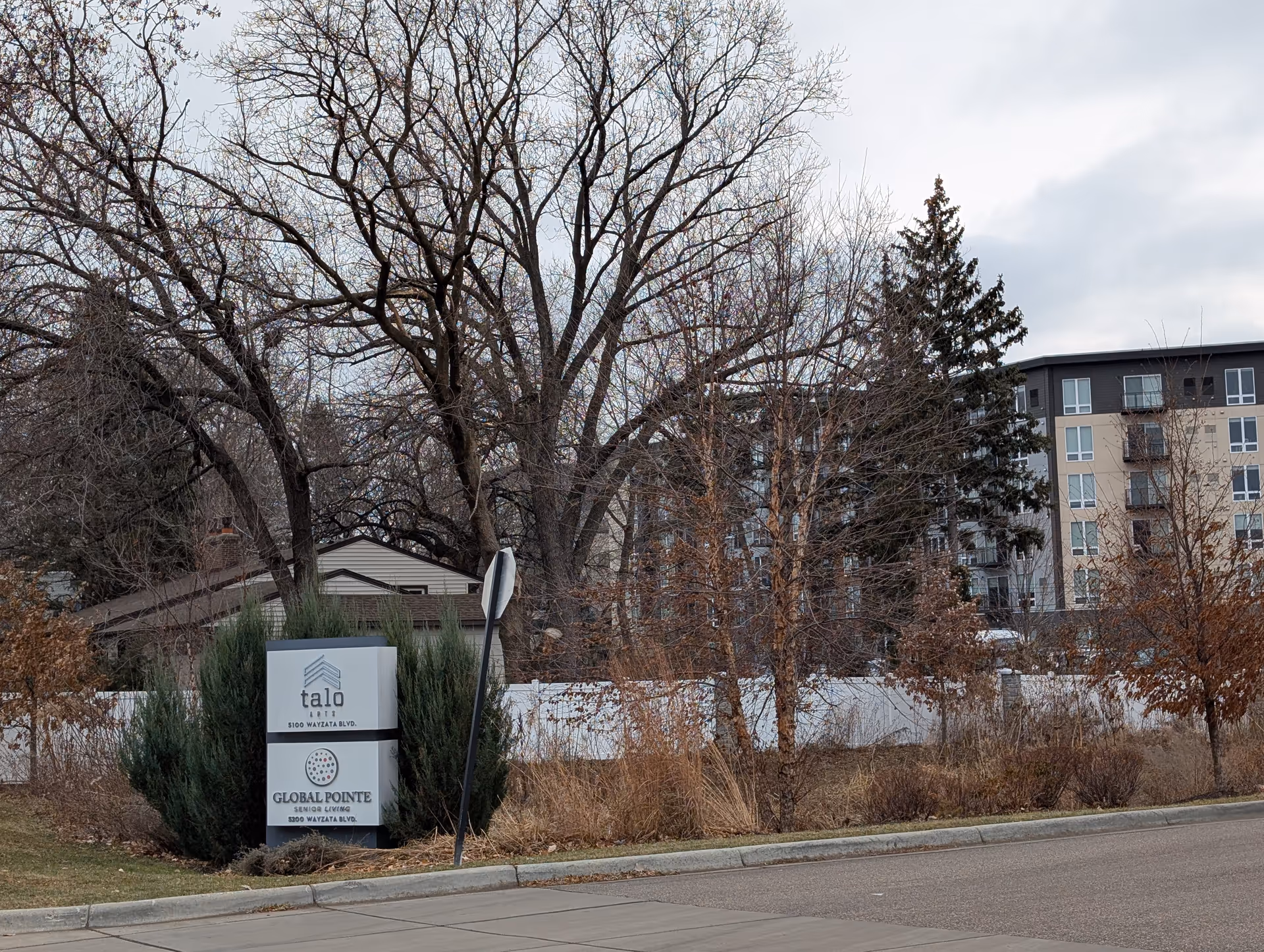 Outdoor view of a street corner with a sign for Global Pointe Senior Living and Talo Apartments, surrounded by leafless trees and bushes, with a multi-story building in the background under a cloudy sky.