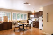A bright kitchen and dining area with wooden floors, a round table with four chairs, dark wood cabinets, a white refrigerator, and a stove. Large windows with white blinds allow natural light to fill the room.
