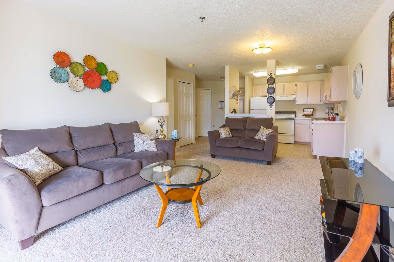 A bright living room with beige carpet, featuring a gray three-seater sofa and a matching loveseat with patterned throw pillows. A round glass coffee table with wooden legs sits in the center. The room opens into a kitchen area with light wood cabinets, a white refrigerator, stove, and countertops. Decorative wall art with colorful circular designs is mounted above the sofa, and a lamp with a white shade is on a side table next to the sofa.