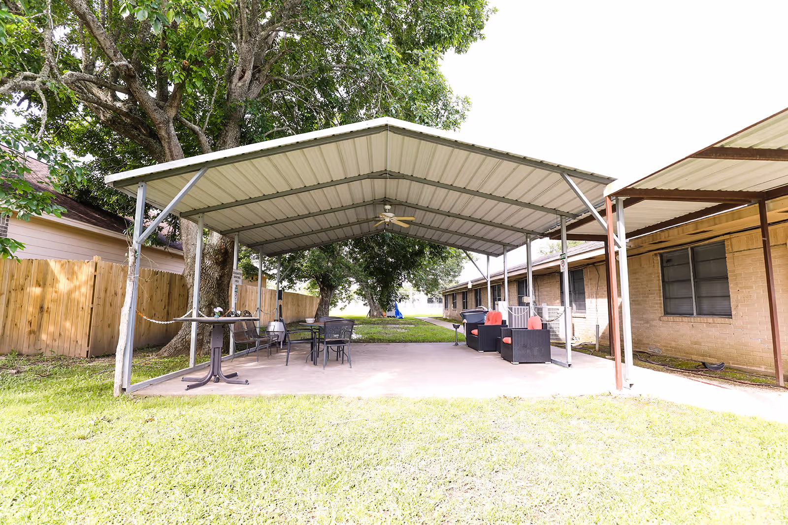 Covered outdoor patio with tables, chairs and lounge seating next to a brick building and grassy yard.