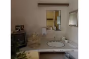A bathroom sink area with a round mirror above it. On the countertop, there is a soap dispenser, a decorative birdcage, and a small television to the left. The walls are light-colored, and there is a framed picture hanging on the right wall.