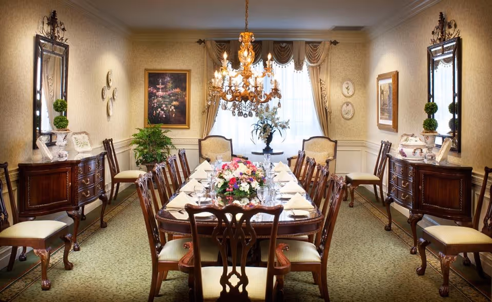 Elegant dining room with a long wooden table set for a meal, surrounded by matching wooden chairs with cushioned seats. The table is decorated with floral centerpieces and neatly folded napkins. The room features two ornate sideboards with mirrors above them, framed artwork on the walls, a large chandelier hanging from the ceiling, and a window with draped curtains letting in natural light.