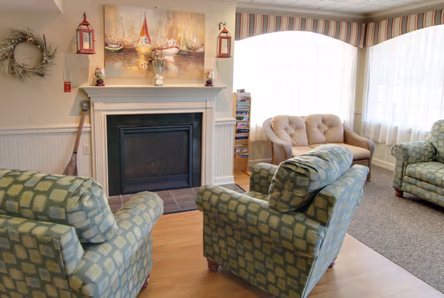 A cozy living room area with a fireplace in the center, flanked by two red lanterns and a painting of boats above the mantel. There are two patterned armchairs facing the fireplace and a cushioned loveseat near a large window with sheer curtains and a striped valance. The room has light-colored walls and a mix of wood and carpet flooring.