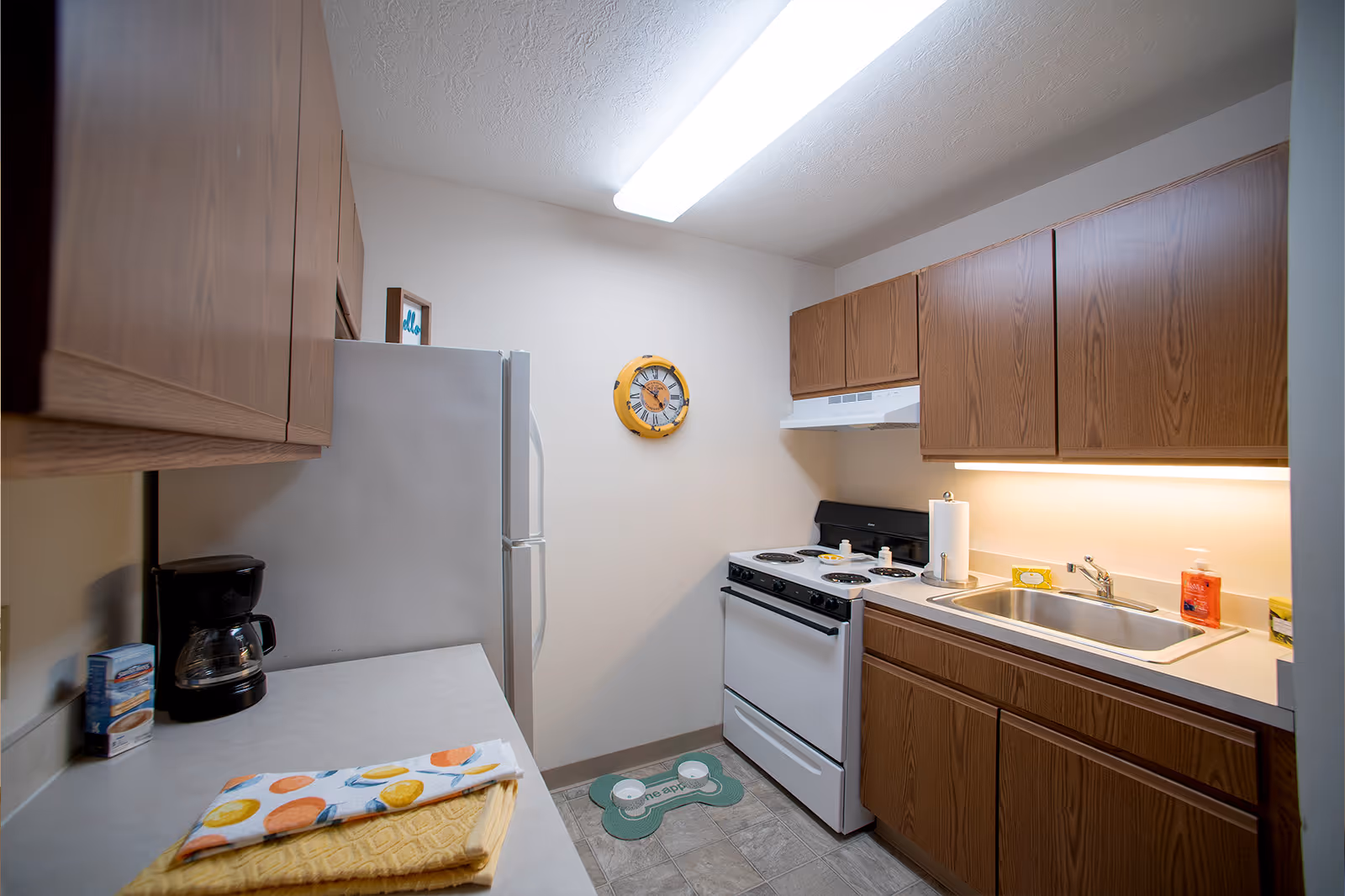 A small kitchen with wooden cabinets, a white refrigerator, a white electric stove with four burners, a stainless steel sink, and a countertop with a coffee maker and folded kitchen towels. A yellow clock is mounted on the wall above the stove, and a fluorescent ceiling light illuminates the space.