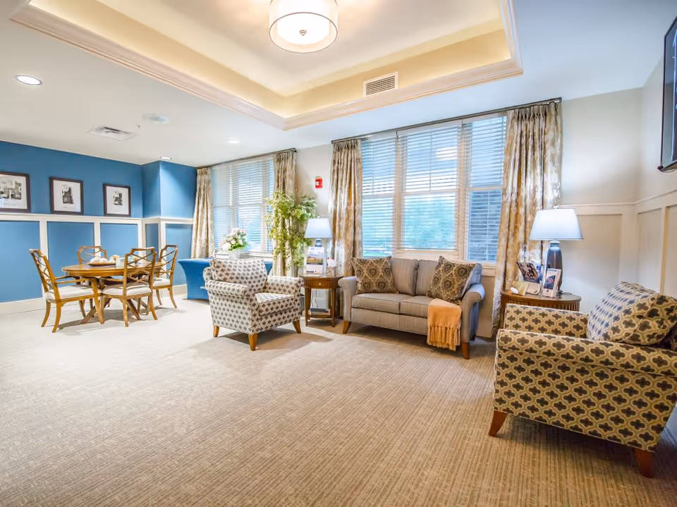 Bright furnished memory-care common room with a sofa, patterned armchairs, a dining table, large windows with blinds and lamps.
