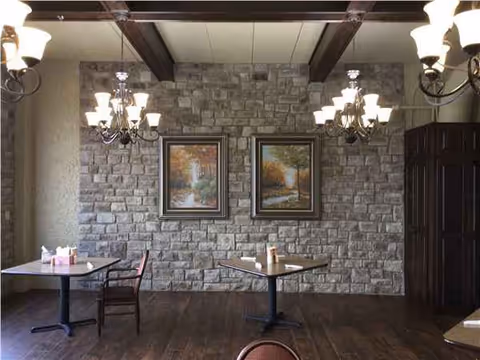 Stone-walled dining area with two tables, chandeliers, and framed landscape paintings.