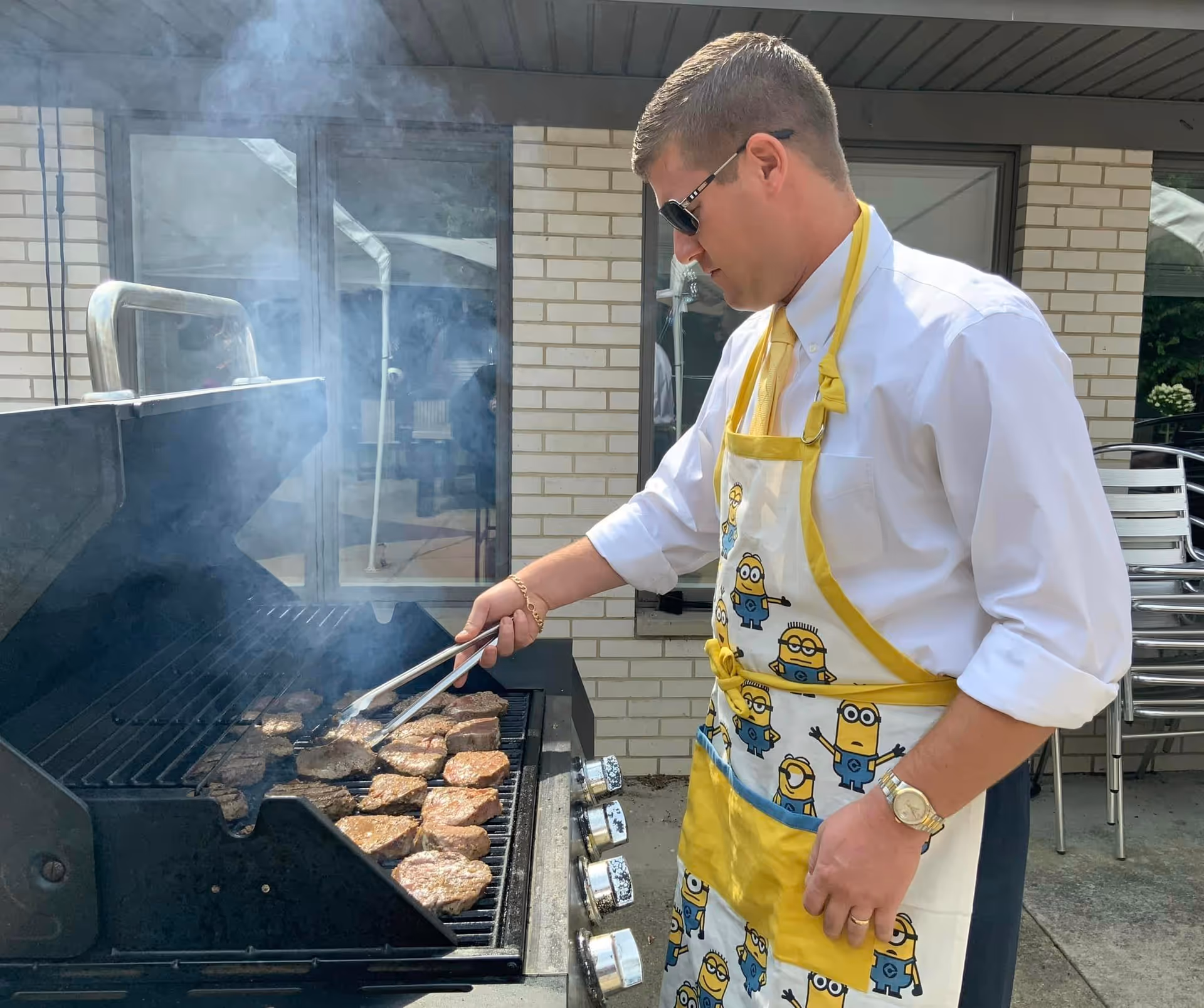 A man wearing sunglasses, a white shirt, and a Minions-themed apron is grilling multiple hamburger patties on an outdoor barbecue grill. The setting appears to be outside a brick building with windows and stacked chairs nearby.