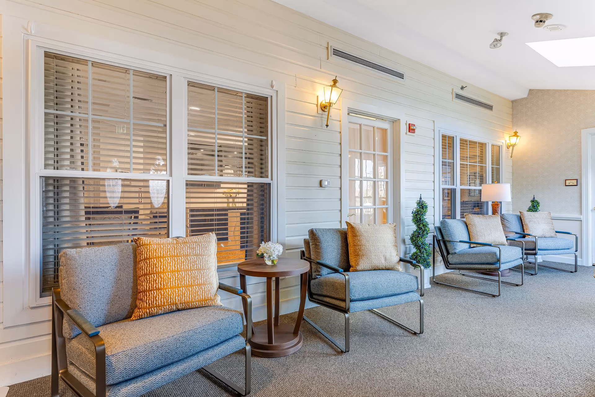 Sunlit seating area with a row of upholstered armchairs, side tables, lamps, and windows along a paneled interior wall.