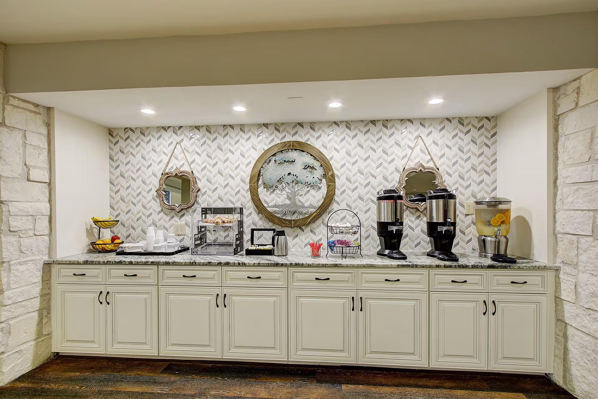 A beverage and snack station with a long white cabinet topped with granite countertop. On the counter are two beverage dispensers, a glass container with lemon water, a tiered fruit basket, disposable cups, and a small display case with snacks. The wall behind features a patterned tile backsplash, two decorative mirrors, and a round wall art piece depicting a tree.