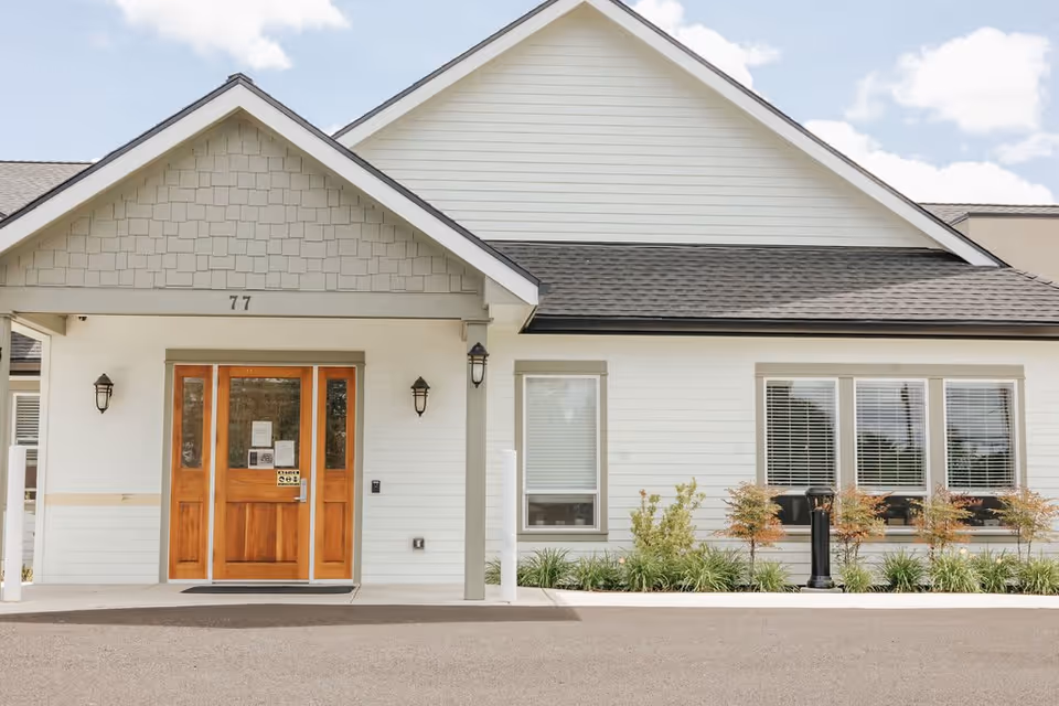 Exterior front view of a single-story building with white siding and a gray shingled roof. The entrance features wooden double doors with glass panels, flanked by two wall-mounted lantern-style lights. There are several windows with white blinds and a small landscaped area with shrubs and plants in front of the building. The number 77 is displayed above the entrance.