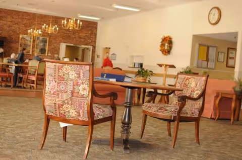 Senior living facility lounge with patterned chairs around tables and residents in background.