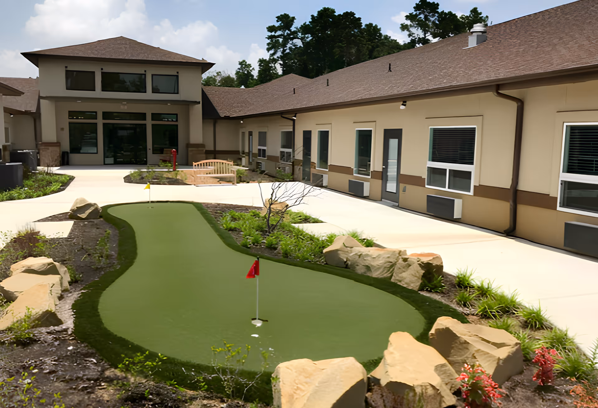 Outdoor courtyard area at Crimson Heights Health & Wellness featuring a small putting green with two red flags, surrounded by landscaped plants and large rocks. The courtyard is bordered by a beige building with multiple windows and doors, and a bench is visible near the center. Trees and a partly cloudy sky are in the background.