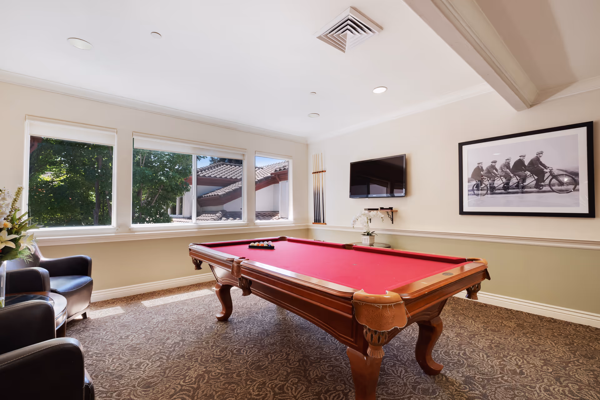 A well-lit game room with a red felt pool table in the center, two black leather chairs to the left, a wall-mounted flat screen TV, a framed black and white photo of five men riding a multi-seat bicycle, and three large windows showing trees and part of a tiled roof outside.