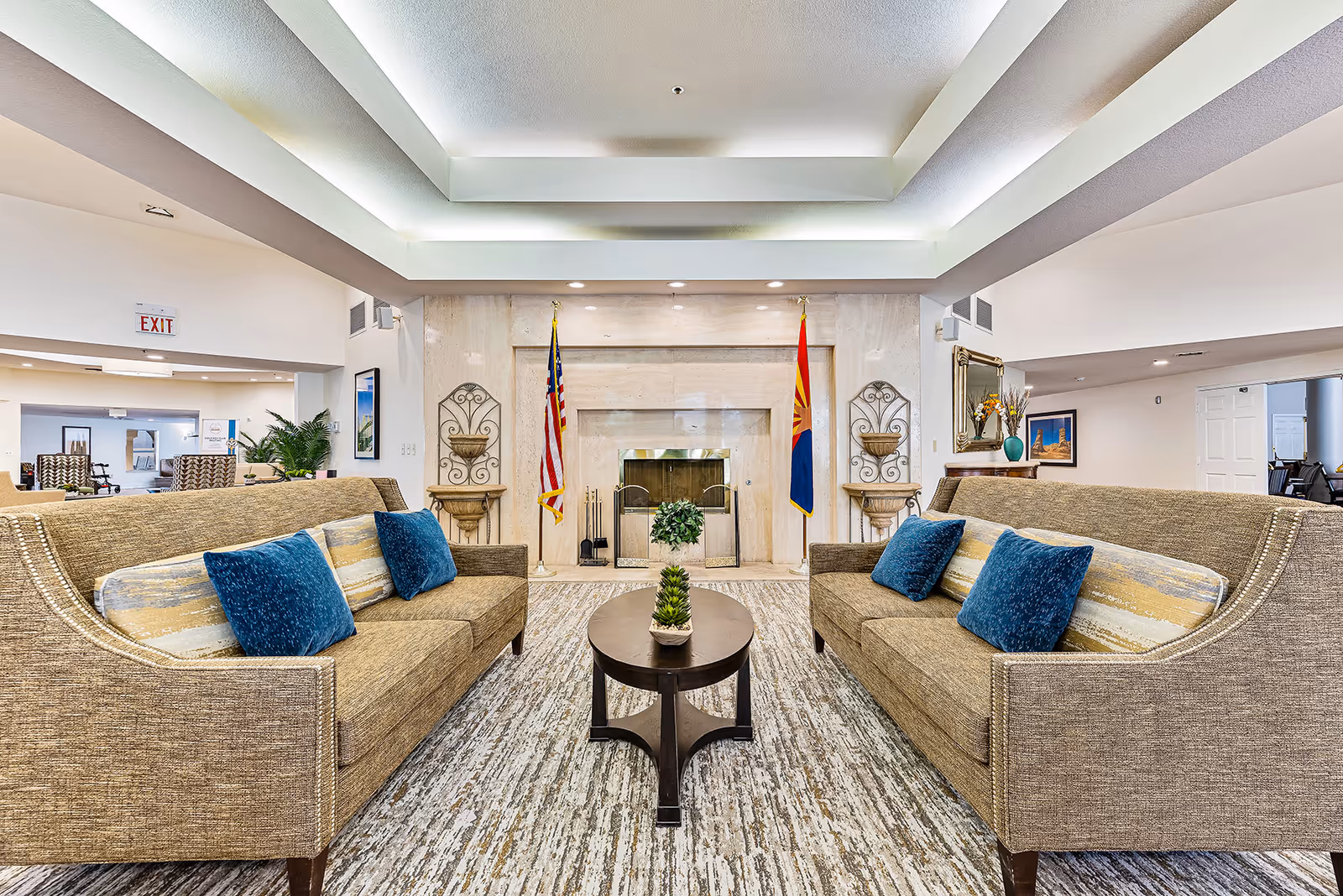 A bright and spacious senior living facility common area featuring two beige sofas with blue and patterned cushions facing each other across a dark wooden coffee table with a small plant. In the background, there is a large marble fireplace flanked by the American flag and the Arizona state flag, with decorative wall sconces on either side. The ceiling has recessed lighting and a tray design, and the floor is covered with a patterned carpet. Additional seating and decor are visible in the surrounding areas.