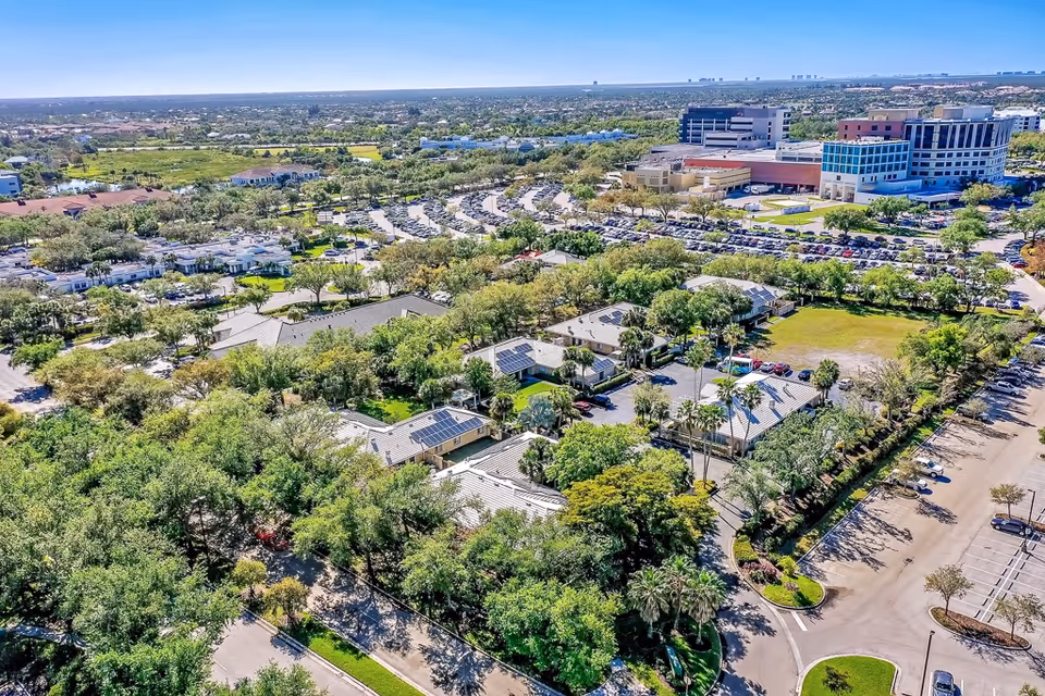 Aerial view of Healthpark Senior Living facility surrounded by trees and greenery, with multiple buildings, parking lots filled with cars, and a distant cityscape under a clear blue sky.