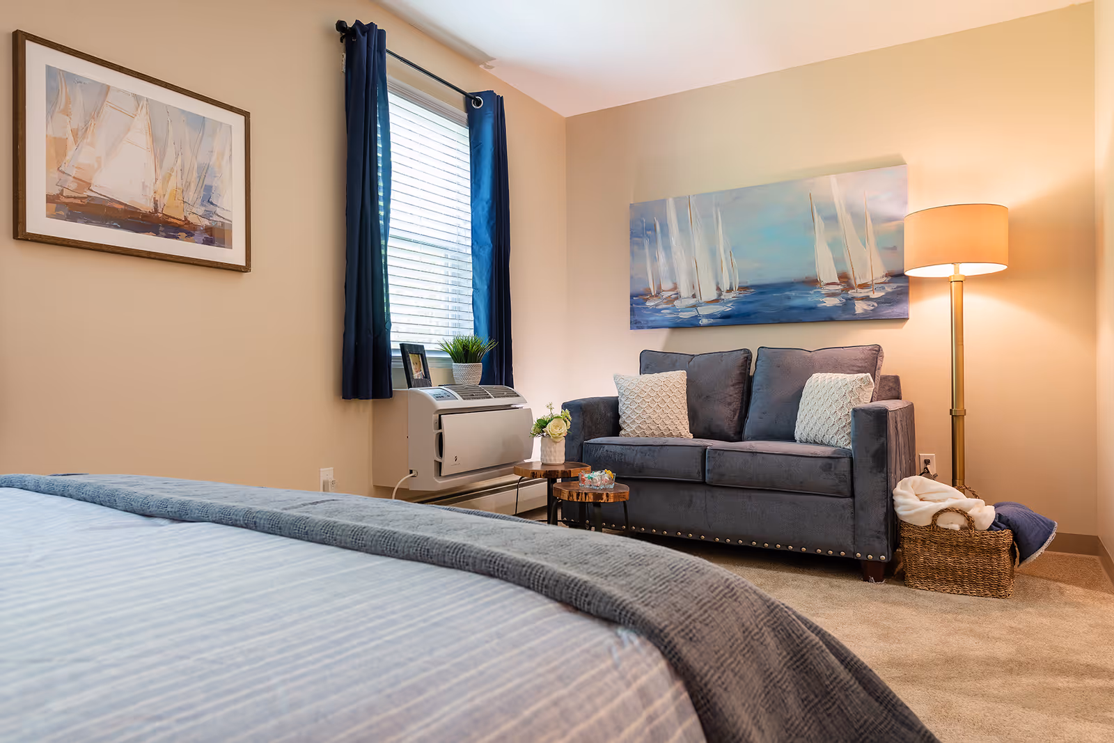 A cozy bedroom featuring a bed with a blue and gray blanket in the foreground, a dark gray loveseat with two patterned pillows against the wall, a floor lamp with a beige shade, a wicker basket with blankets, a window with dark blue curtains, and two paintings of sailboats on the walls.