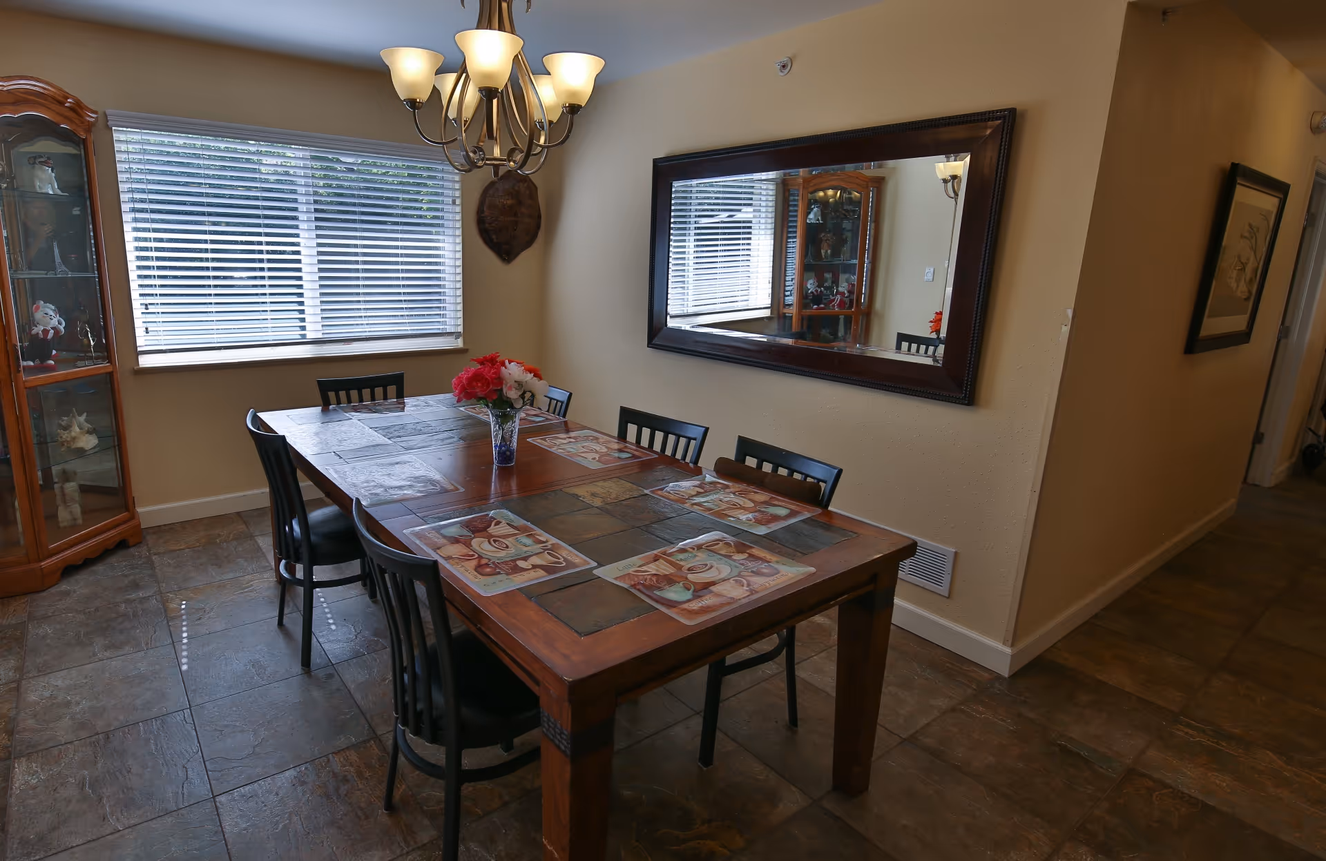 A dining room with a rectangular wooden table set with six black chairs. The table has placemats and a vase with red and white flowers in the center. A large mirror hangs on the wall to the right, reflecting the window with closed blinds on the left. A chandelier with multiple lights hangs above the table. There is a wooden display cabinet in the corner near the window and a framed picture on the wall in the hallway to the right.