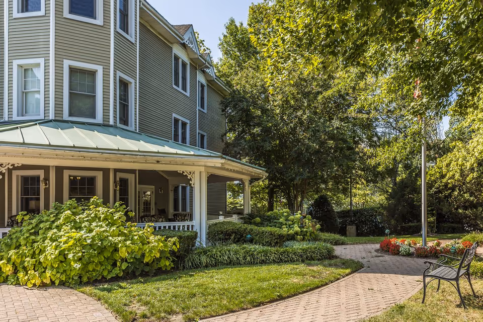 Exterior view of a multi-story senior living facility with a covered porch, surrounded by lush green trees, bushes, and a well-maintained garden. A paved walkway curves through the garden, with a bench and a flagpole with flowers around its base.