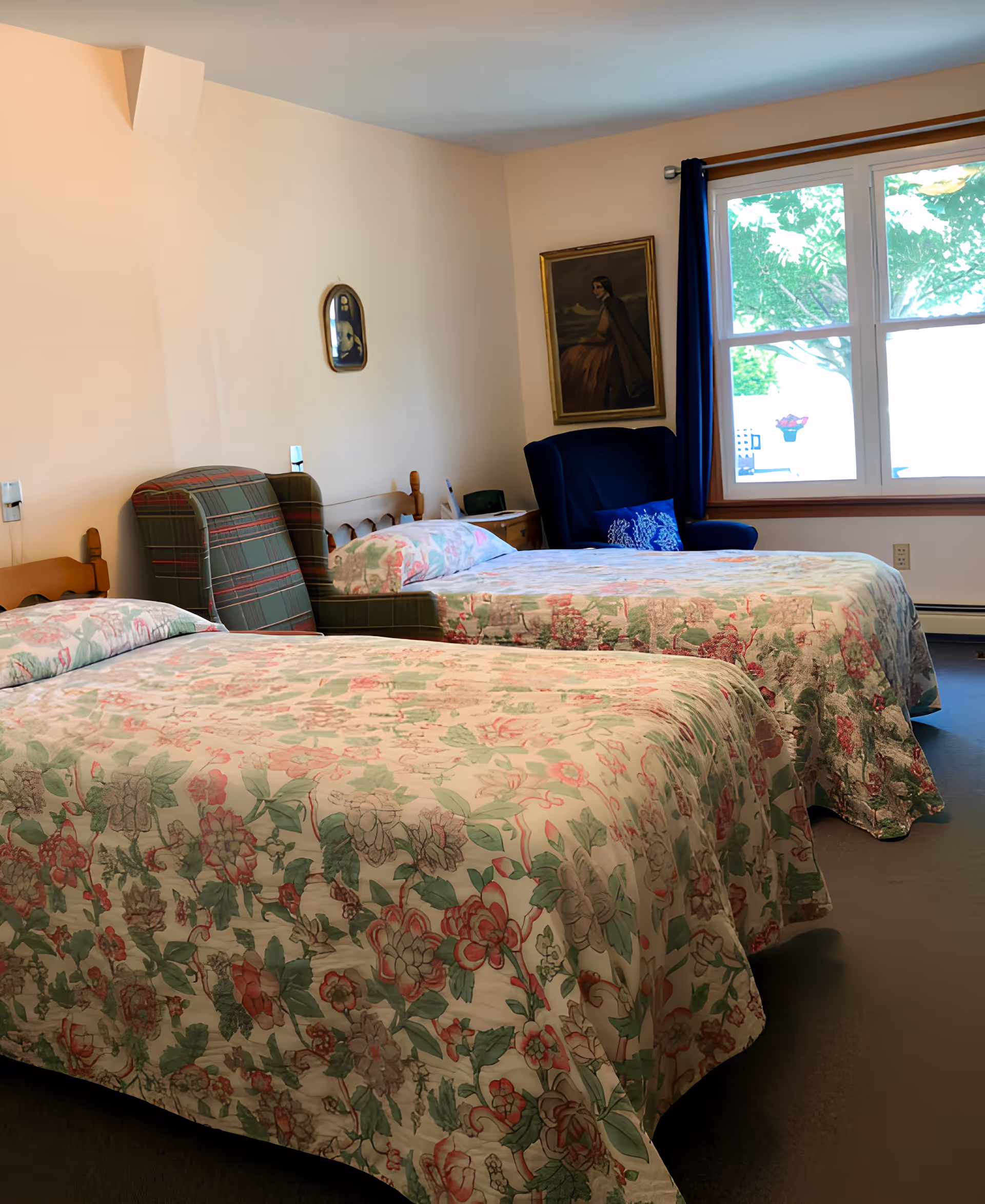 A bedroom with two twin beds covered in floral bedspreads. There is a green plaid armchair between the beds and a blue armchair near a window with blue curtains. A framed painting hangs on the wall above the blue armchair.