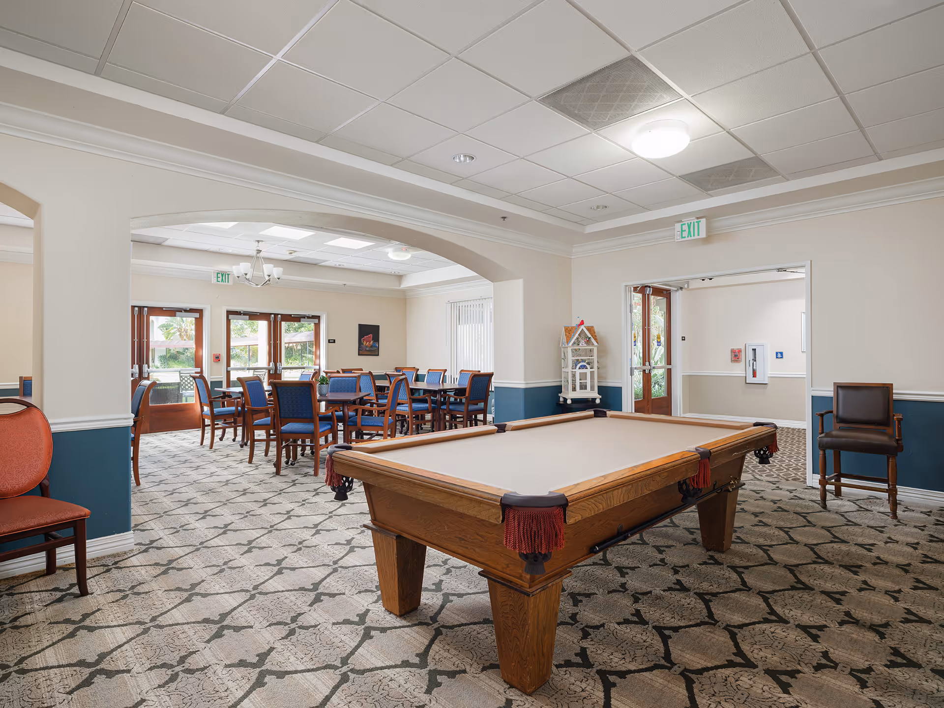Interior view of a senior living facility common area featuring a pool table in the foreground and a dining area with tables and chairs in the background. The room has patterned carpet, white walls with blue wainscoting, and multiple exit doors with windows letting in natural light.