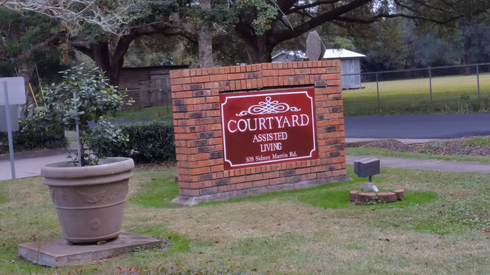 A brick sign on a grassy area with a large potted plant nearby. The sign reads 'COURTYARD ASSISTED LIVING 308 Sidney Martin Rd.' Trees and a fenced area are visible in the background.