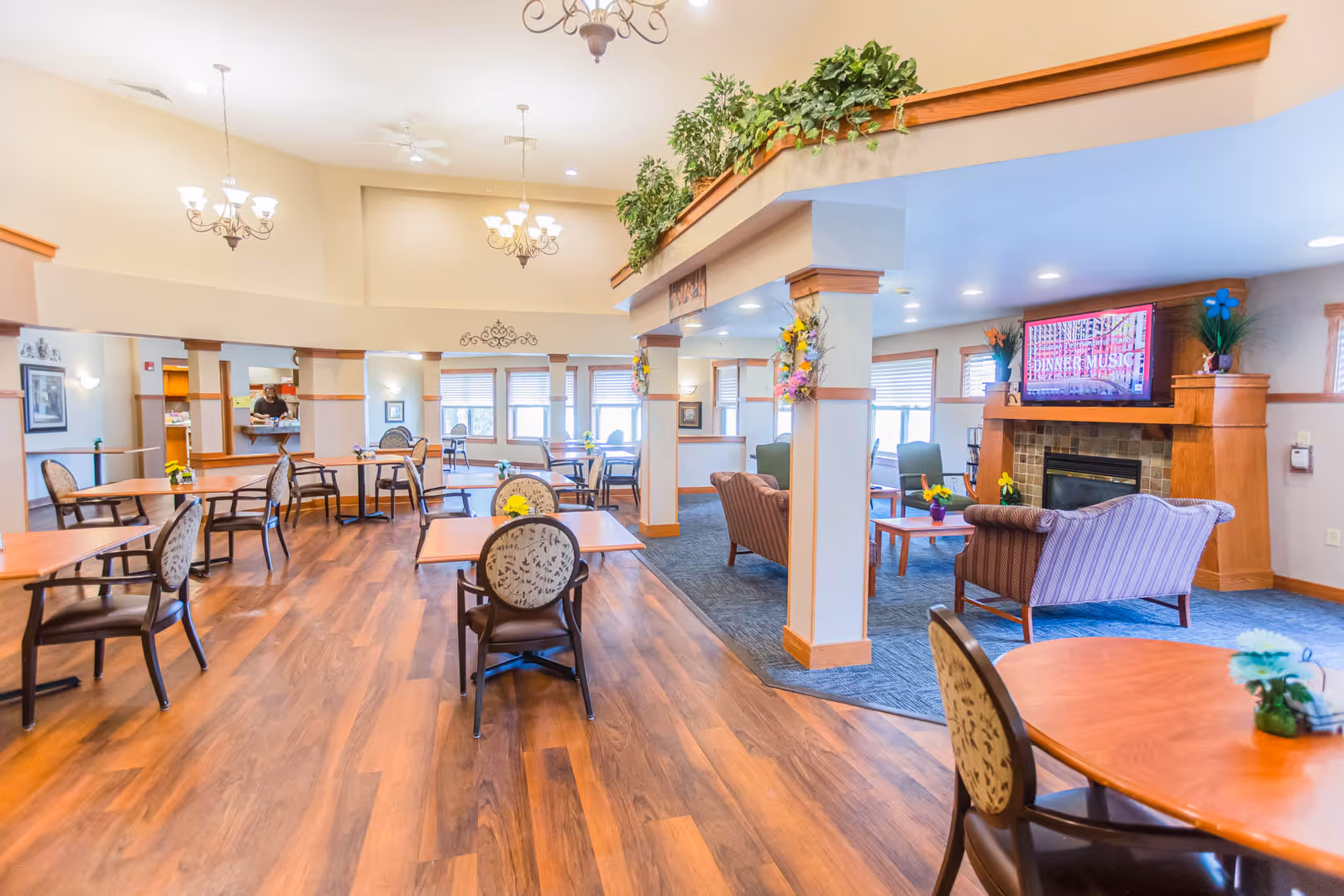 A spacious senior living facility common area with wooden floors and multiple round tables with chairs arranged for dining. The room features high ceilings with chandeliers, large windows allowing natural light, and a cozy seating area with armchairs and a fireplace under a mounted television. Green plants and floral decorations adorn the space, creating a warm and inviting atmosphere.