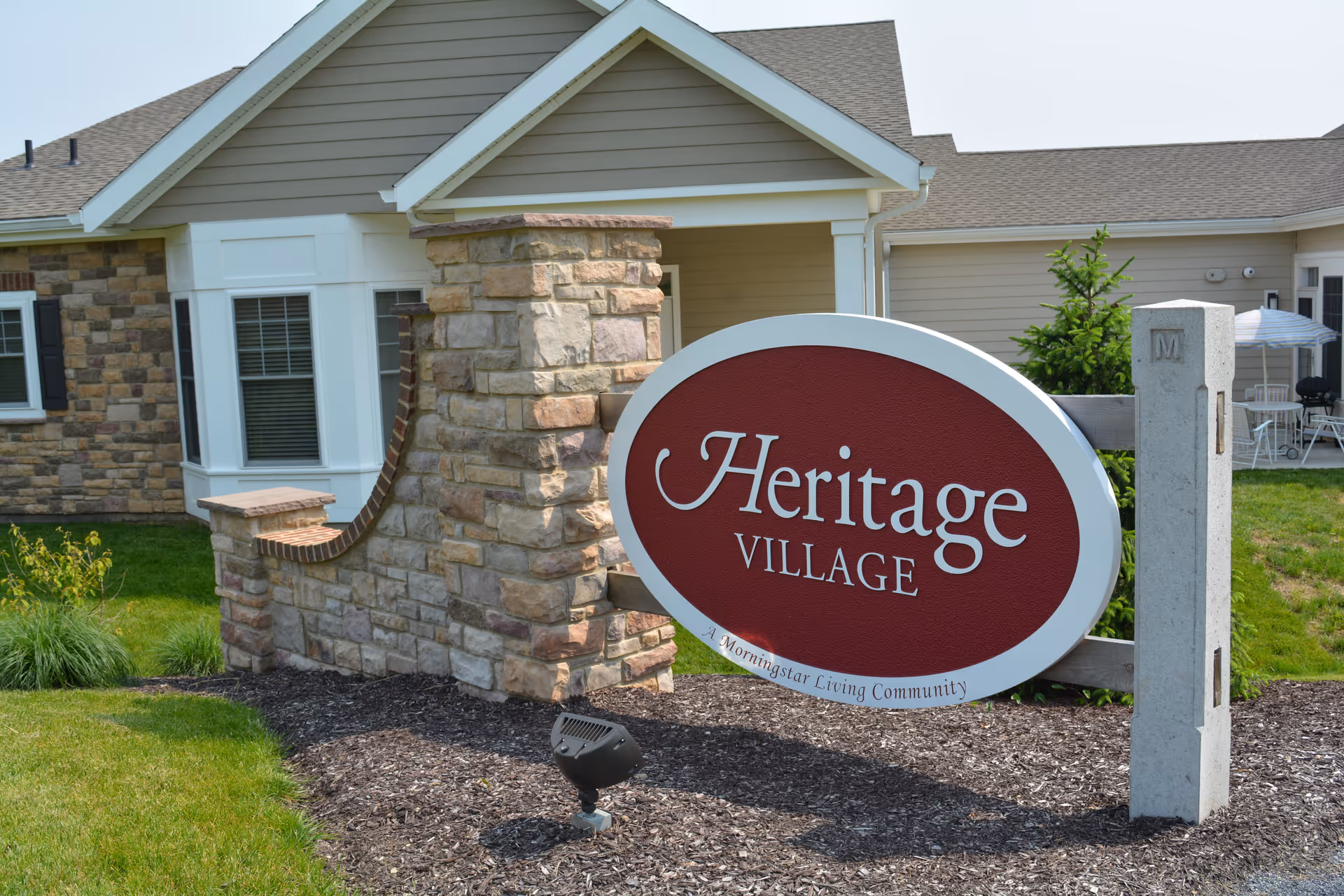 Outdoor view of the entrance sign for Heritage Village, a Morningstar Living Community, with a stone and brick wall and part of a residential building in the background.