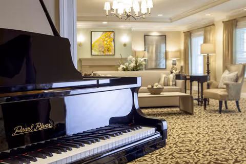 A spacious and elegant living room area featuring a black Pearl River grand piano in the foreground. The room has patterned carpet flooring, beige walls, and large windows with curtains allowing natural light. There is a chandelier hanging from the ceiling, a sofa with cushions, a coffee table with decorative items, an armchair, and framed artwork on the walls.