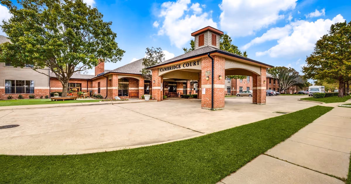 Exterior view of Cambridge Court Senior Living facility showing a brick entrance canopy with the facility name on it, surrounded by trees, a driveway, parked cars, and a sidewalk under a partly cloudy sky.