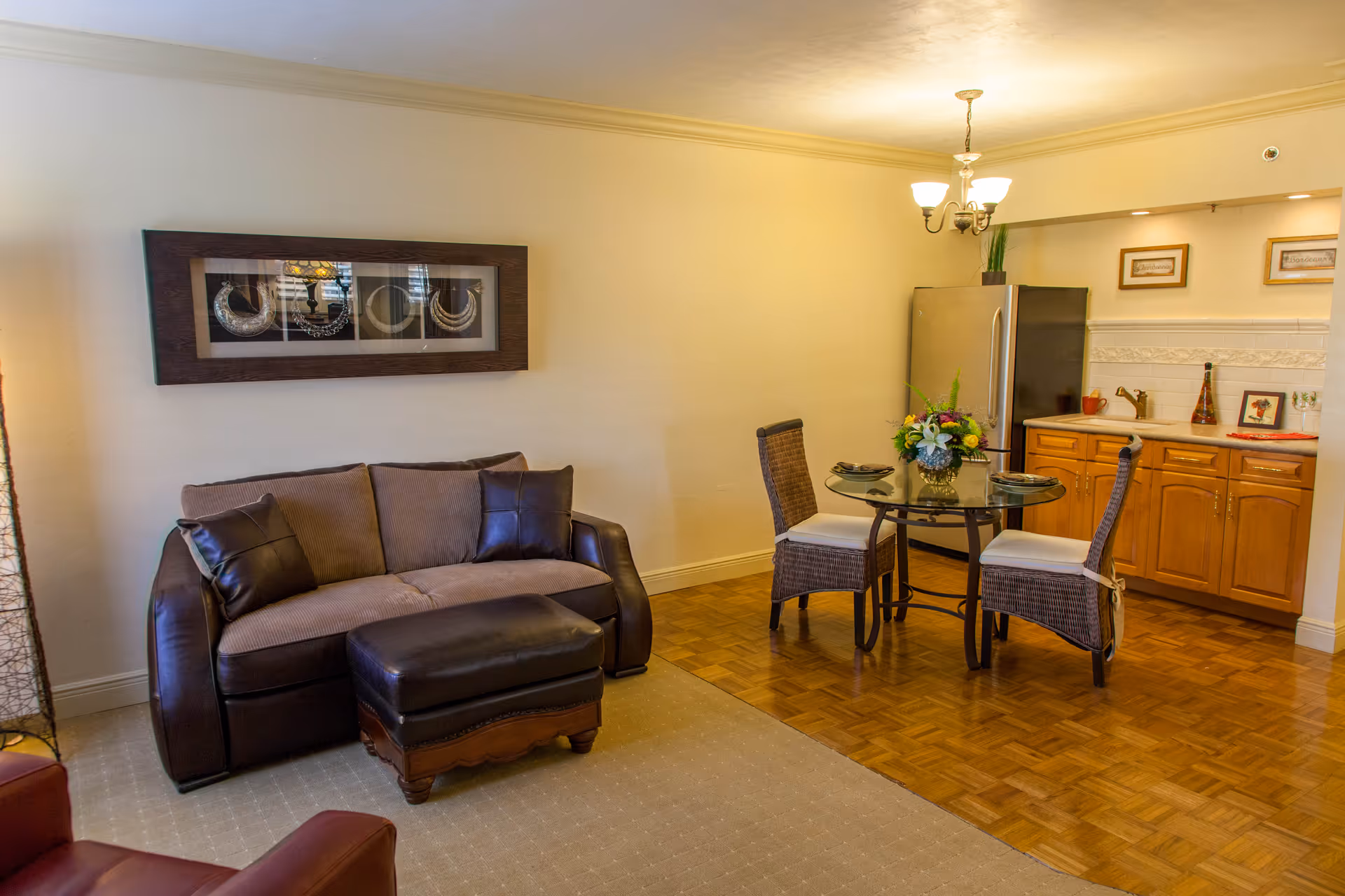 A cozy living area with a brown sofa and matching ottoman on a beige carpet. Adjacent to the living space is a small dining area with a round glass table and two wicker chairs with white cushions. The dining area is next to a kitchenette with wooden cabinets, a stainless steel refrigerator, a sink, and decorative items on the countertop. A chandelier light fixture hangs above the dining table, and a framed artwork is mounted on the wall above the sofa.