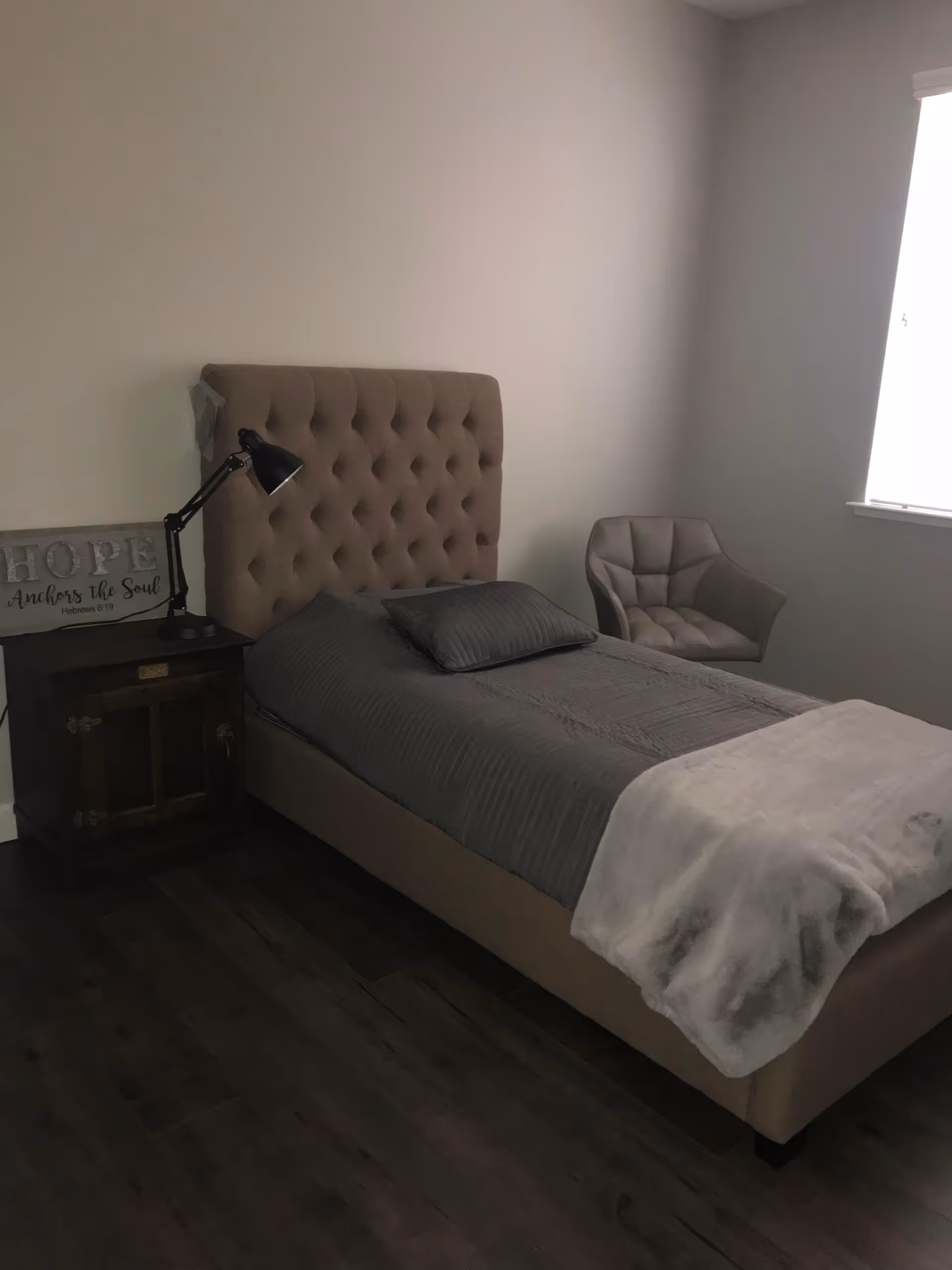 A bedroom with a single bed featuring a tufted beige headboard, gray bedding, and a light gray throw blanket. Next to the bed is a dark wooden nightstand with a black adjustable desk lamp and a decorative sign that reads 'HOPE Anchors the Soul Hebrews 6:19'. A cushioned gray armchair is positioned near a window with white blinds, and the floor is dark wood.