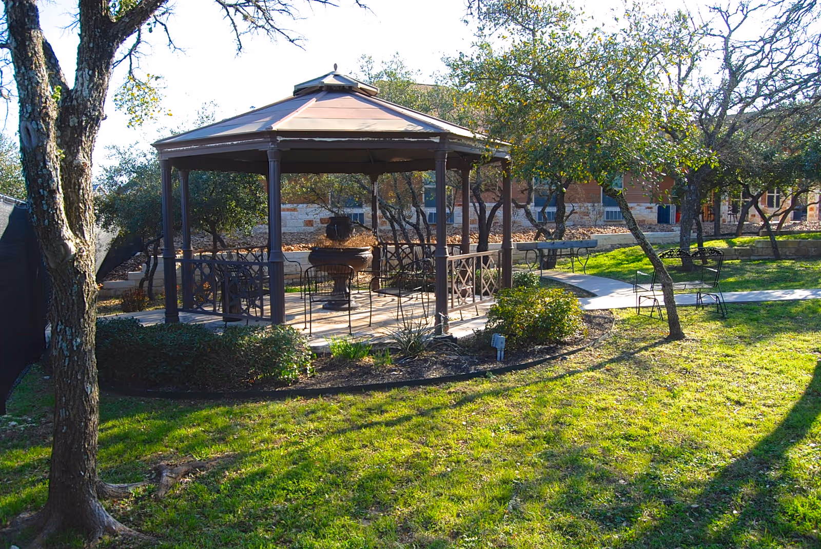 Outdoor garden area with a gazebo featuring a metal roof and several chairs inside. Surrounding the gazebo are trees, bushes, and a well-maintained grassy lawn with a paved walkway leading to the gazebo. A building is visible in the background.