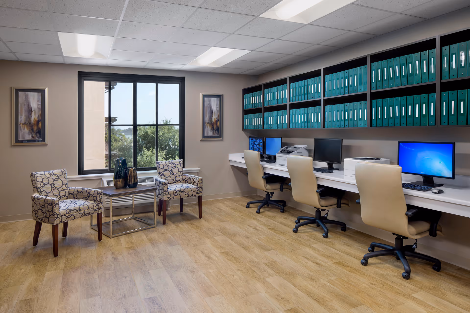 A well-lit office area with three beige swivel chairs in front of a long white desk holding three computer monitors, a printer, and a scanner. Above the desk are shelves filled with green binders. To the left, there are two patterned armchairs with a small table between them holding decorative vases. A large window with a black frame shows greenery outside. Two framed abstract paintings hang on the wall beside the window.