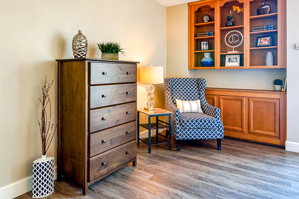 A cozy corner of a room featuring a wooden chest of drawers with decorative items on top, a patterned blue armchair with a small pillow, a side table with a lamp, and built-in wooden shelves and cabinets on the wall displaying various decorative objects and framed photos.