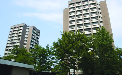 Two tall apartment buildings with multiple windows surrounded by green trees under a clear sky.