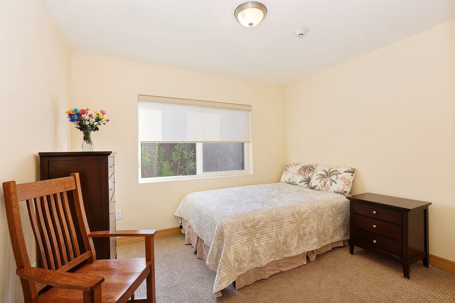 A simple bedroom with a bed covered in a beige quilt and two floral pillows. There is a wooden chair in the foreground, a dark wooden dresser with a vase of colorful flowers on top, and a matching dark wooden nightstand next to the bed. A window with a white roller shade is on the wall behind the bed, and the walls are painted a light cream color.